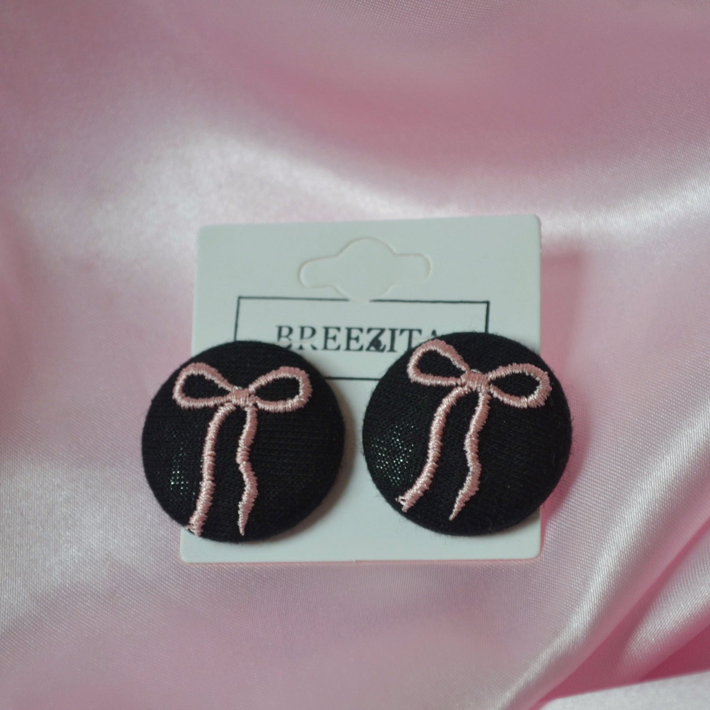 Black earrings with pink bows on a Breezita branded card against a pink satin background