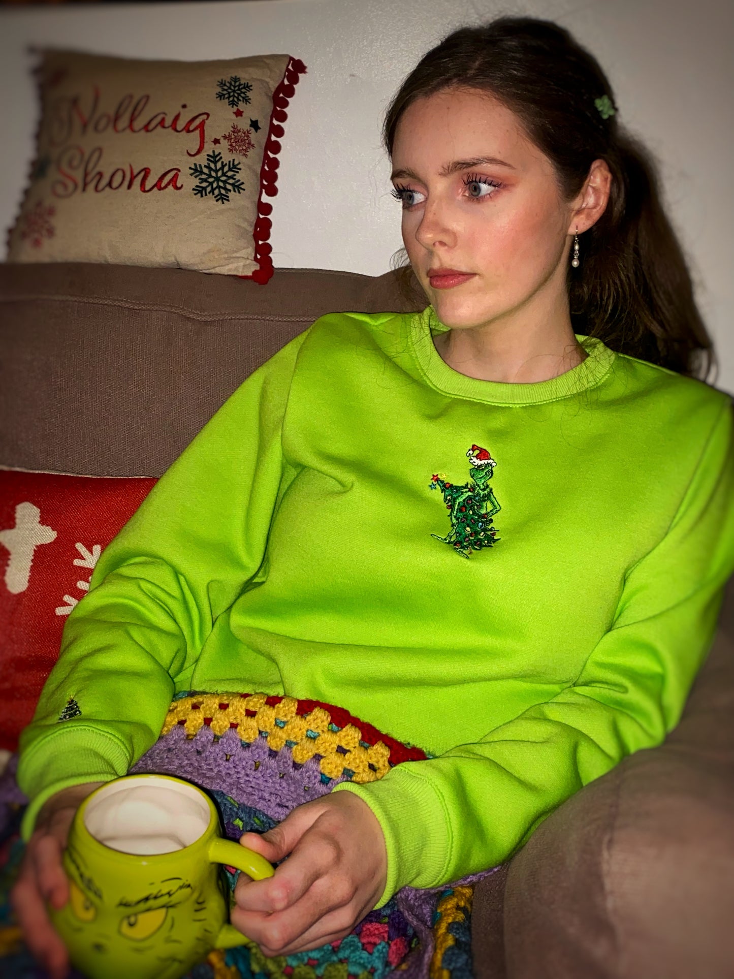 Woman relaxing on sofa in lime-green Christmas sweatshirt with mischievous green hand embroidery, holding matching green mug.