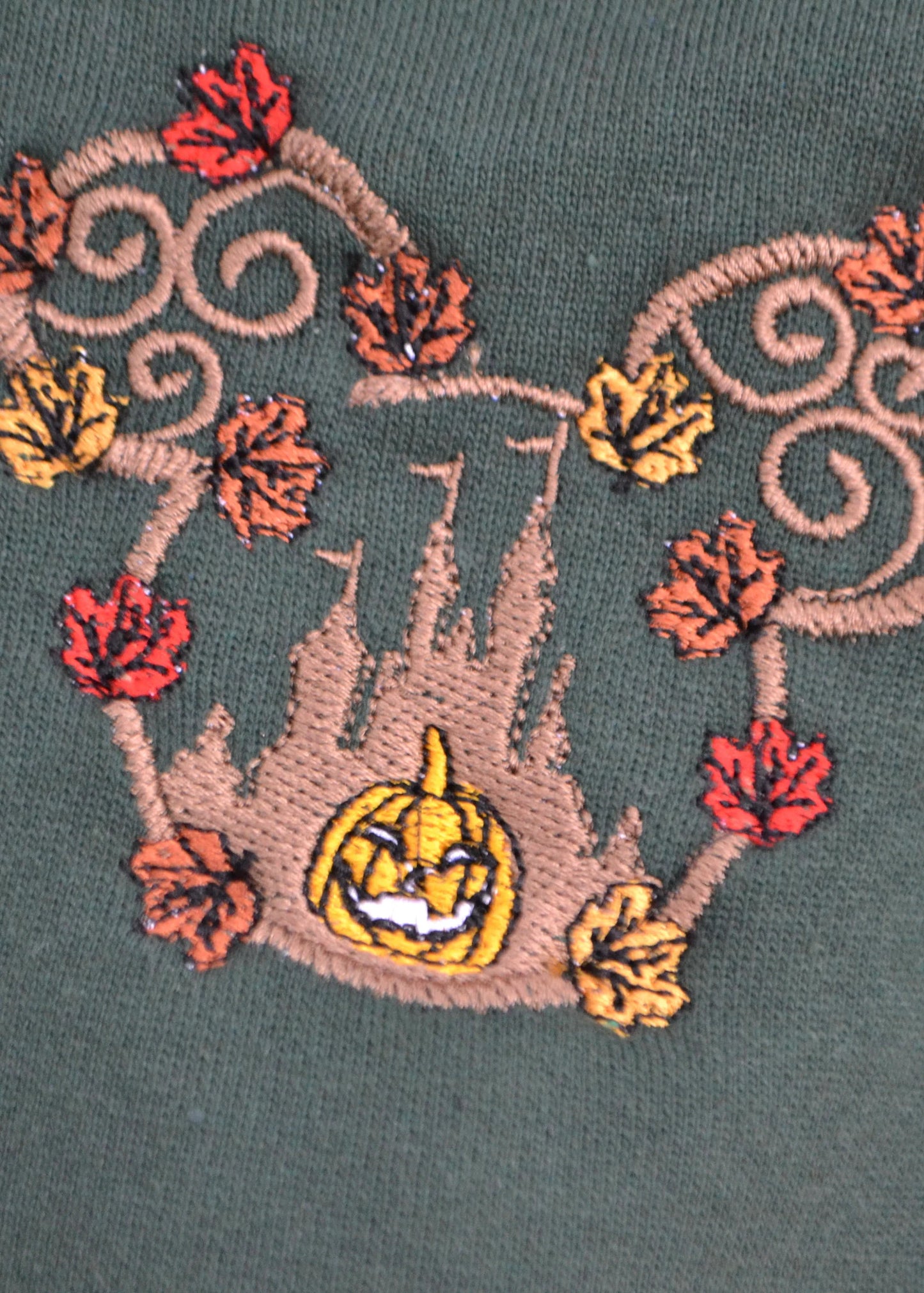 Close-up of autumn pumpkin castle embroidery with swirling vines and colourful leaves on soft forest-green fleece jumper
