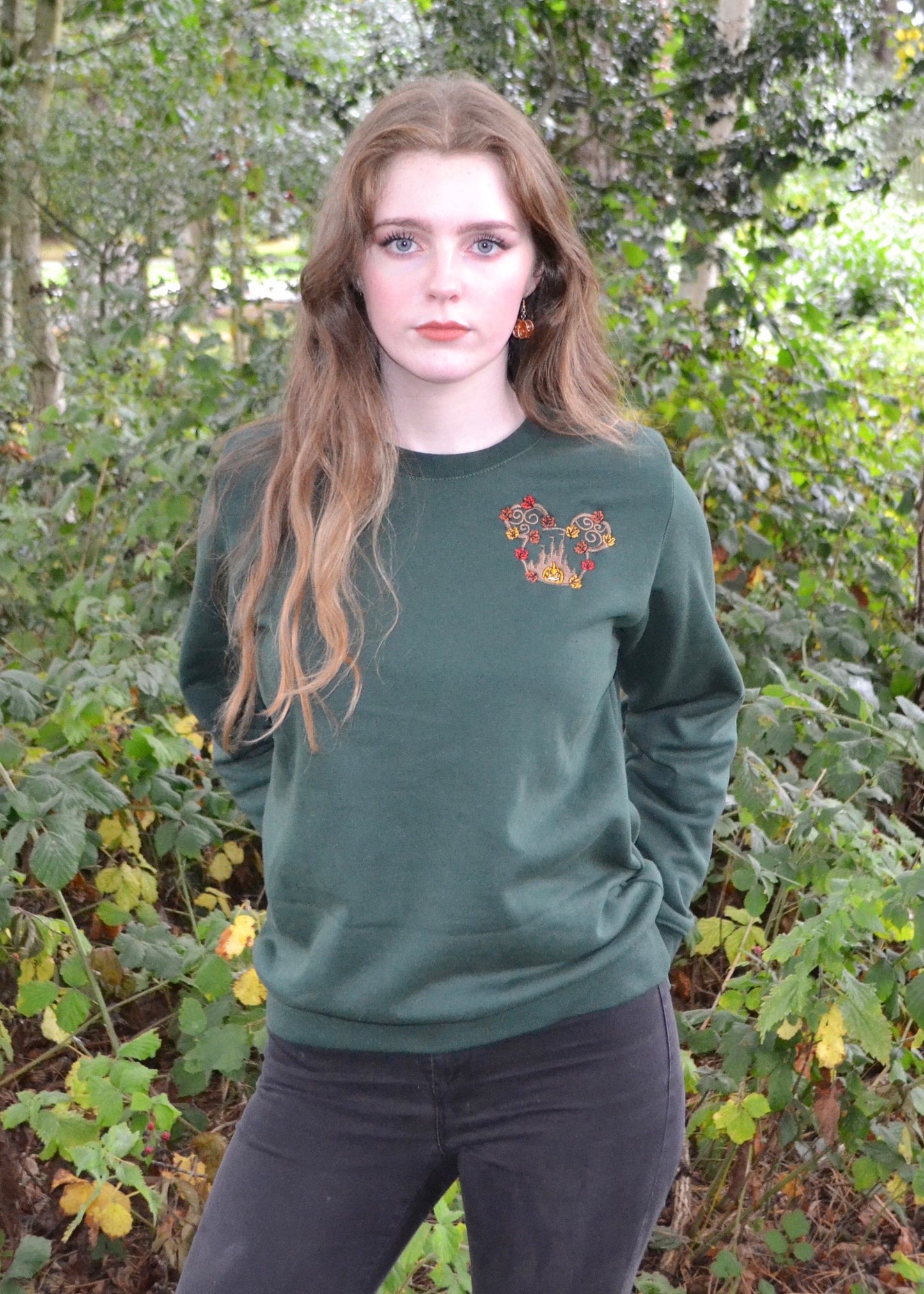 Front view: bottle-green unisex sweatshirt on female model, embroidered pumpkin & swirling leaves, relaxed outdoor setting