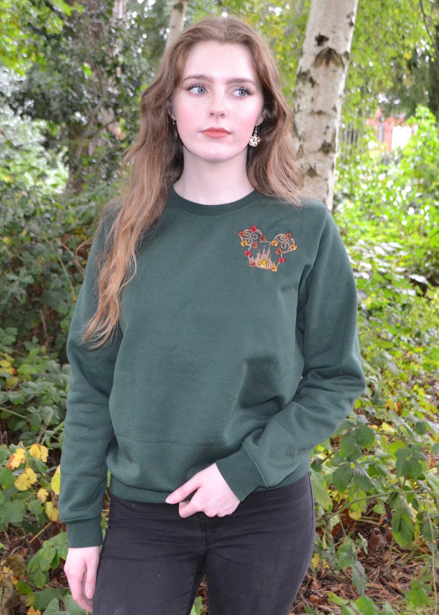 Female model styling bottle-green sweatshirt and black jeans; woodland pumpkin-leaf chest motif, cosy autumn streetwear