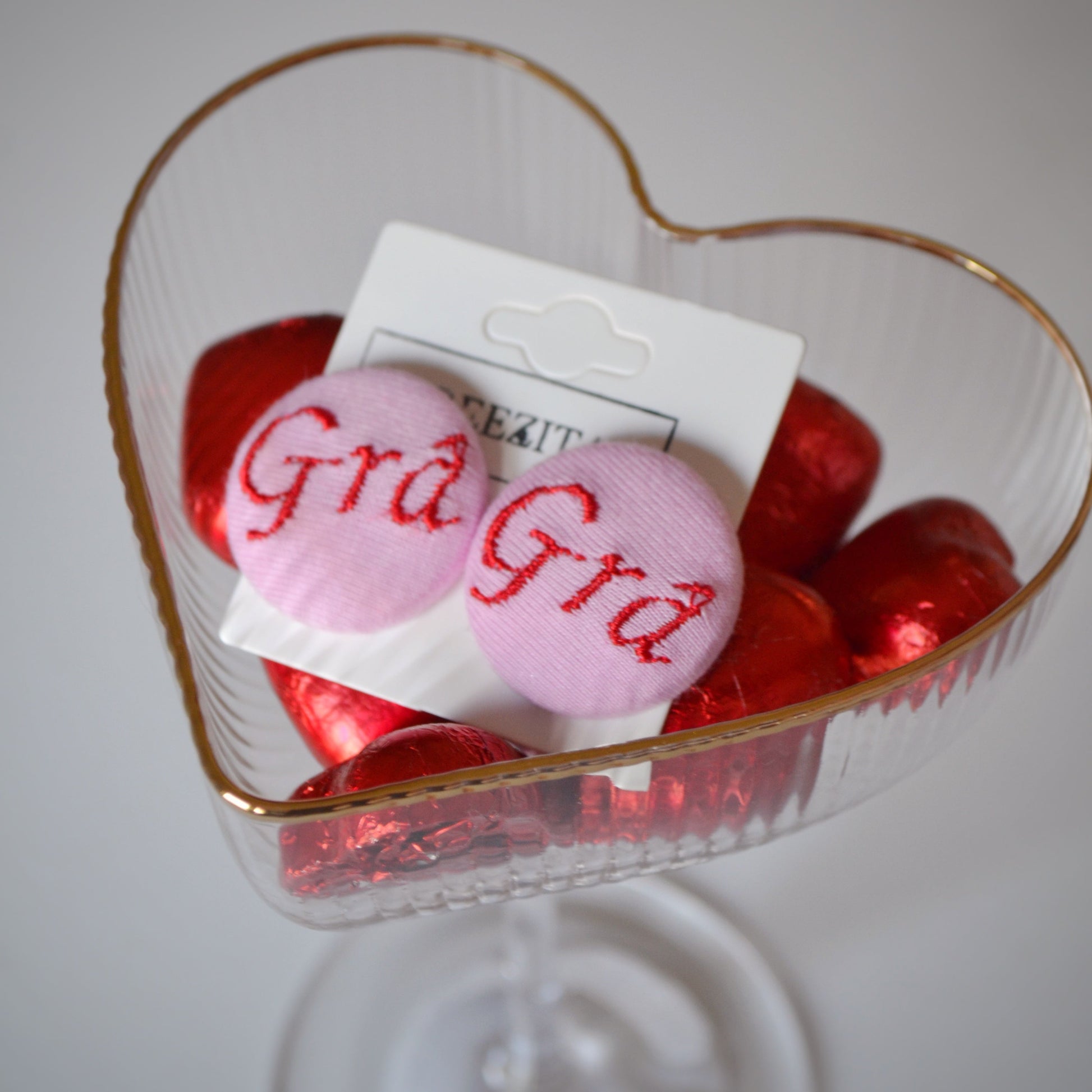 Heart-shaped dish with pink and red Irish button earrings labeled 'Gra' on a white background