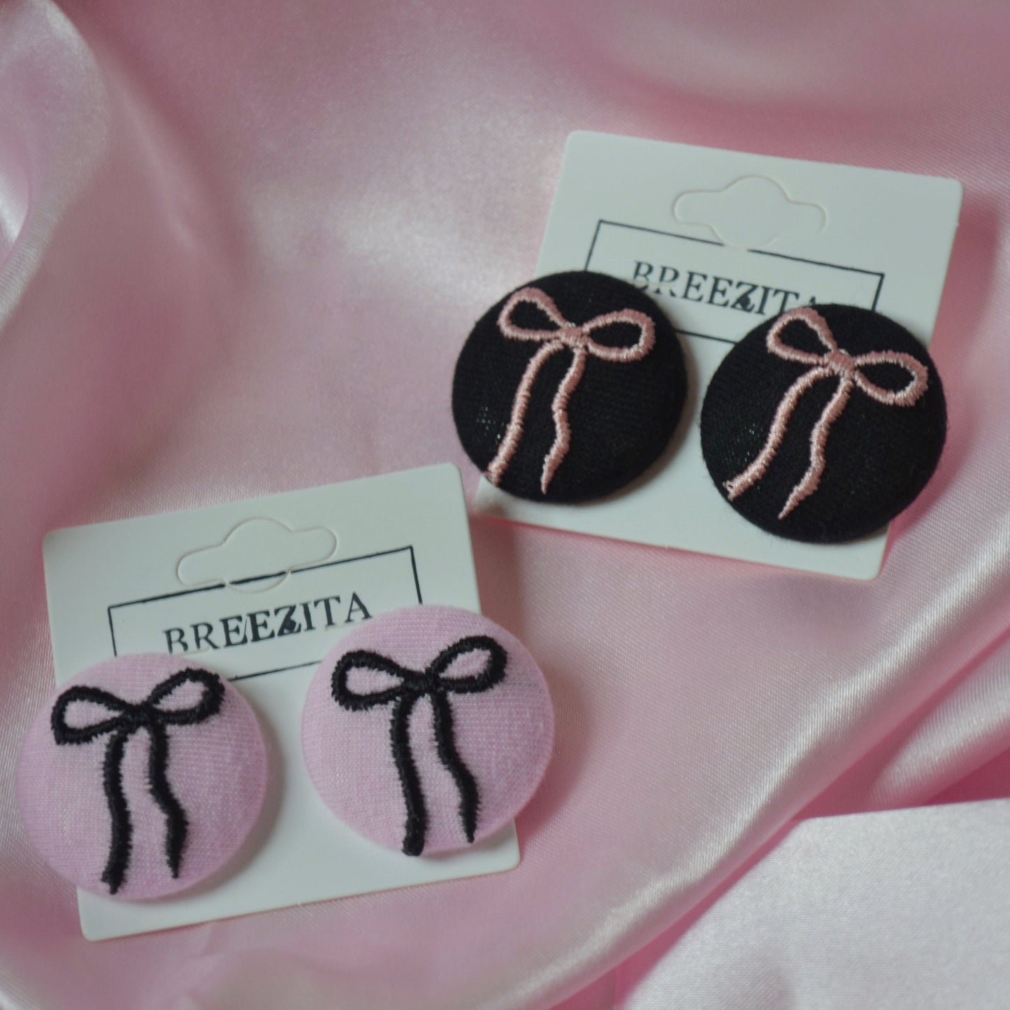 Pair of earrings with pink and black bows on white cards with 'BREEZITA' branding, against a pink satin background.