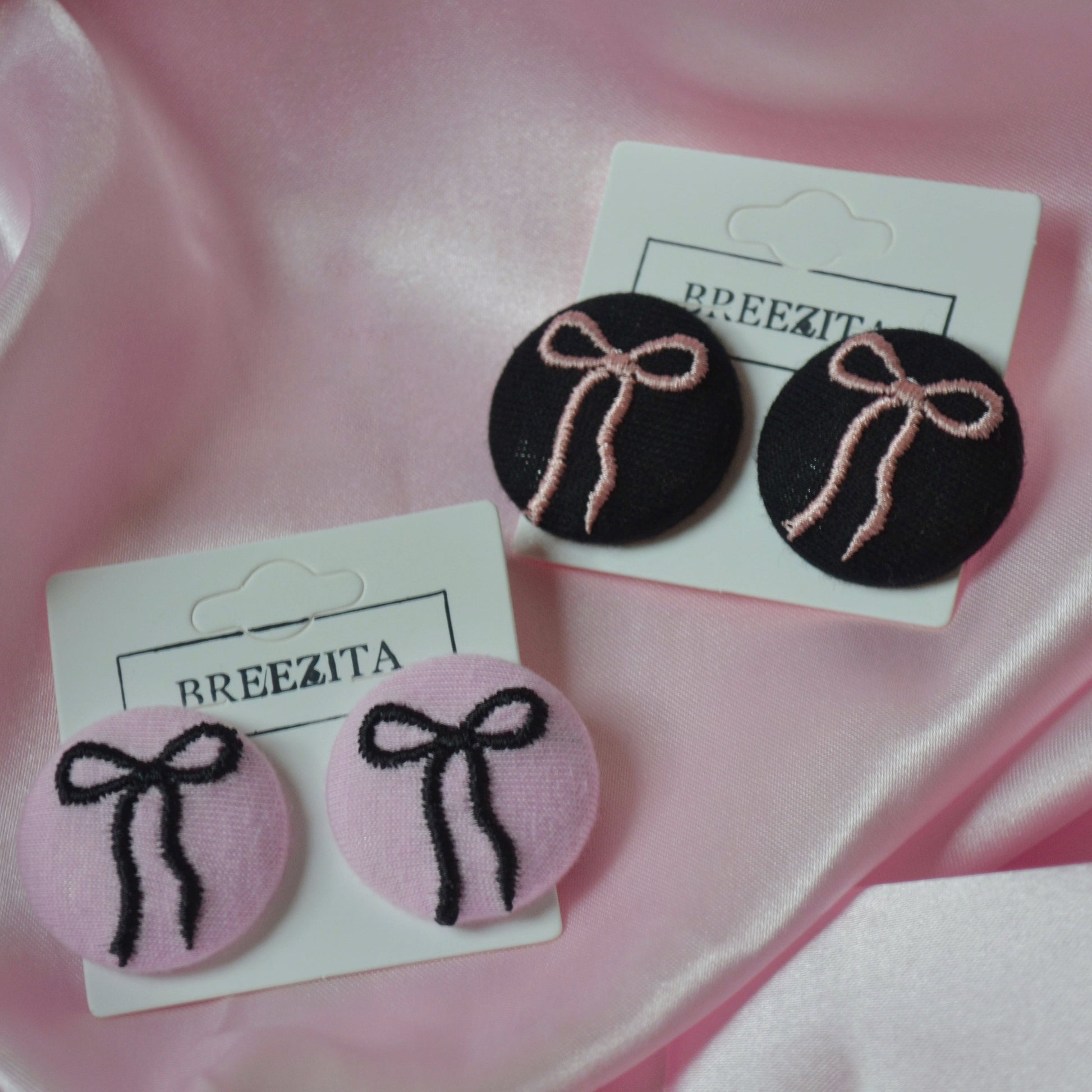 Pair of earrings with pink and black bows on white cards with 'BREEZITA' branding, against a pink satin background.