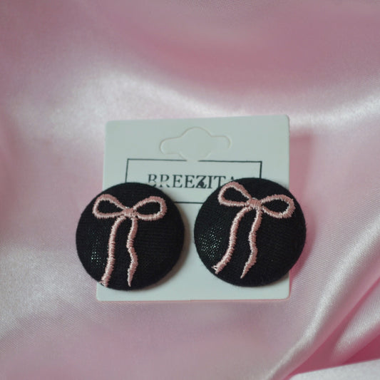 Black earrings with pink bows on a Breezita branded card against a pink satin background