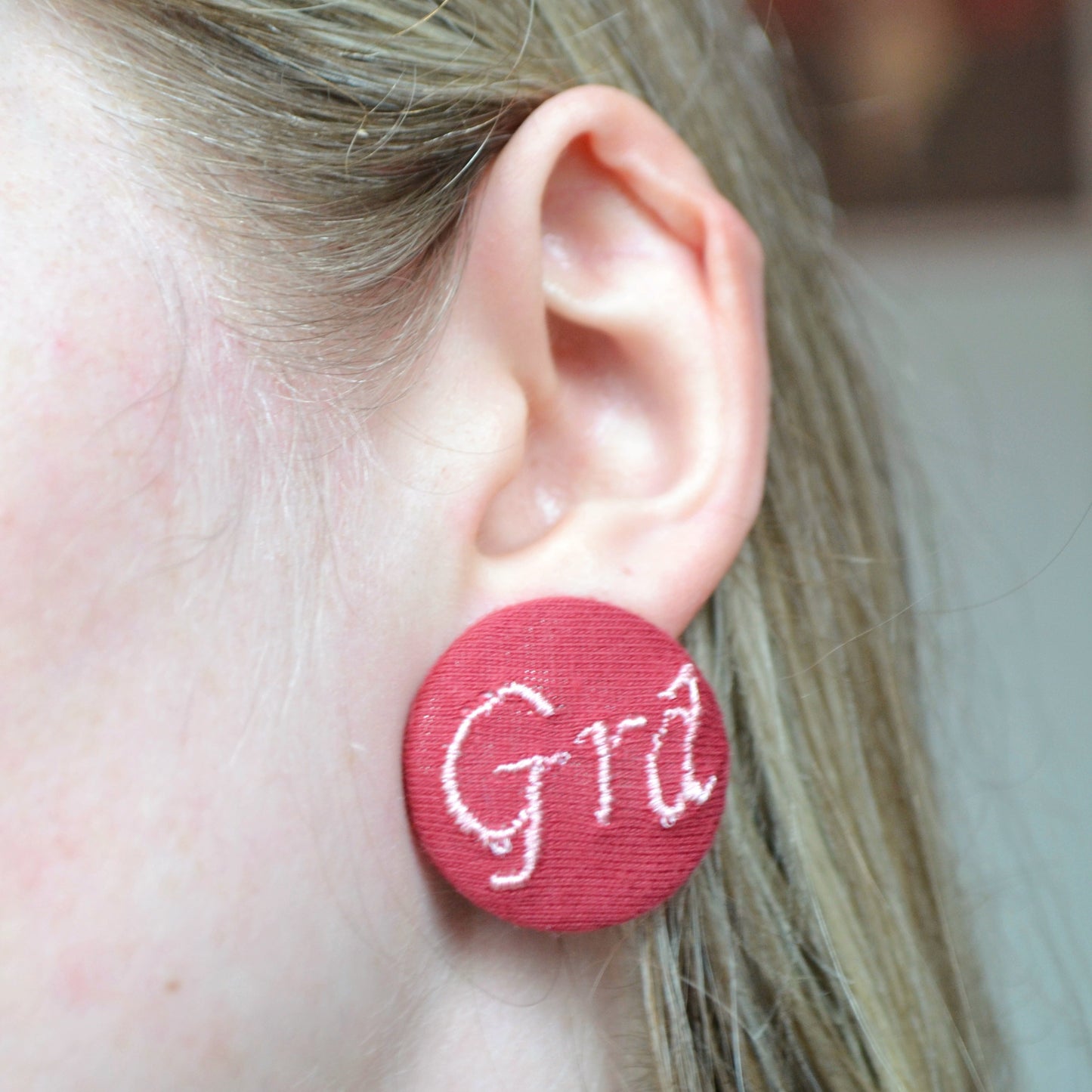Maroon earrings with 'Gra' text worn by a person with blurred background
