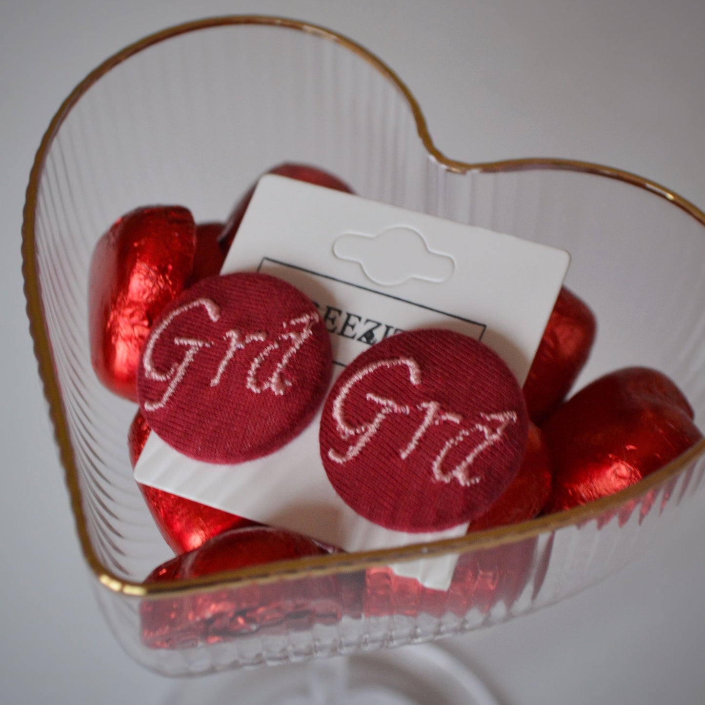 Red button earrings with 'Grá' text in a clear container on a white background