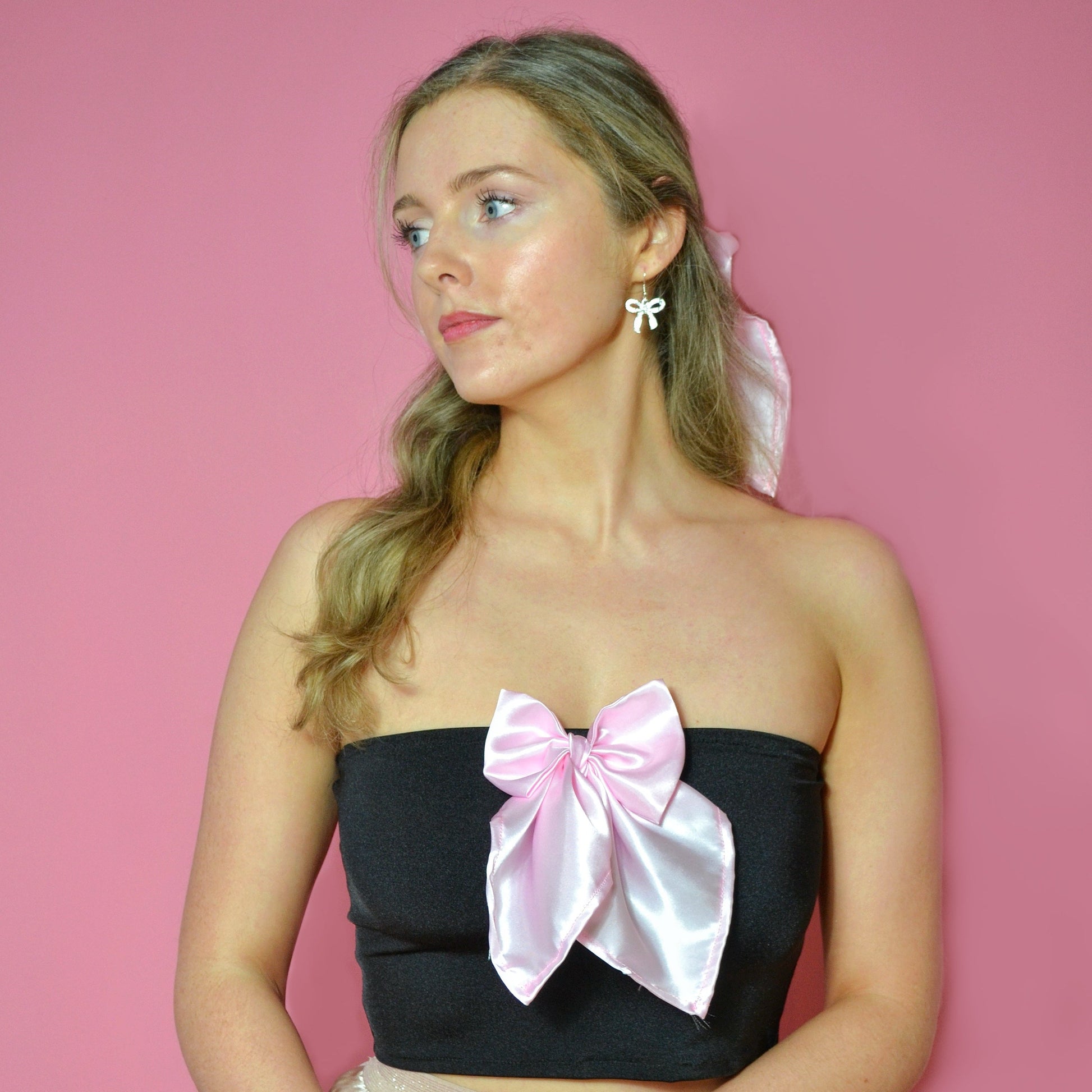 Woman wearing a black strapless top with a large pink bow and a pale pink skirt against a pink background