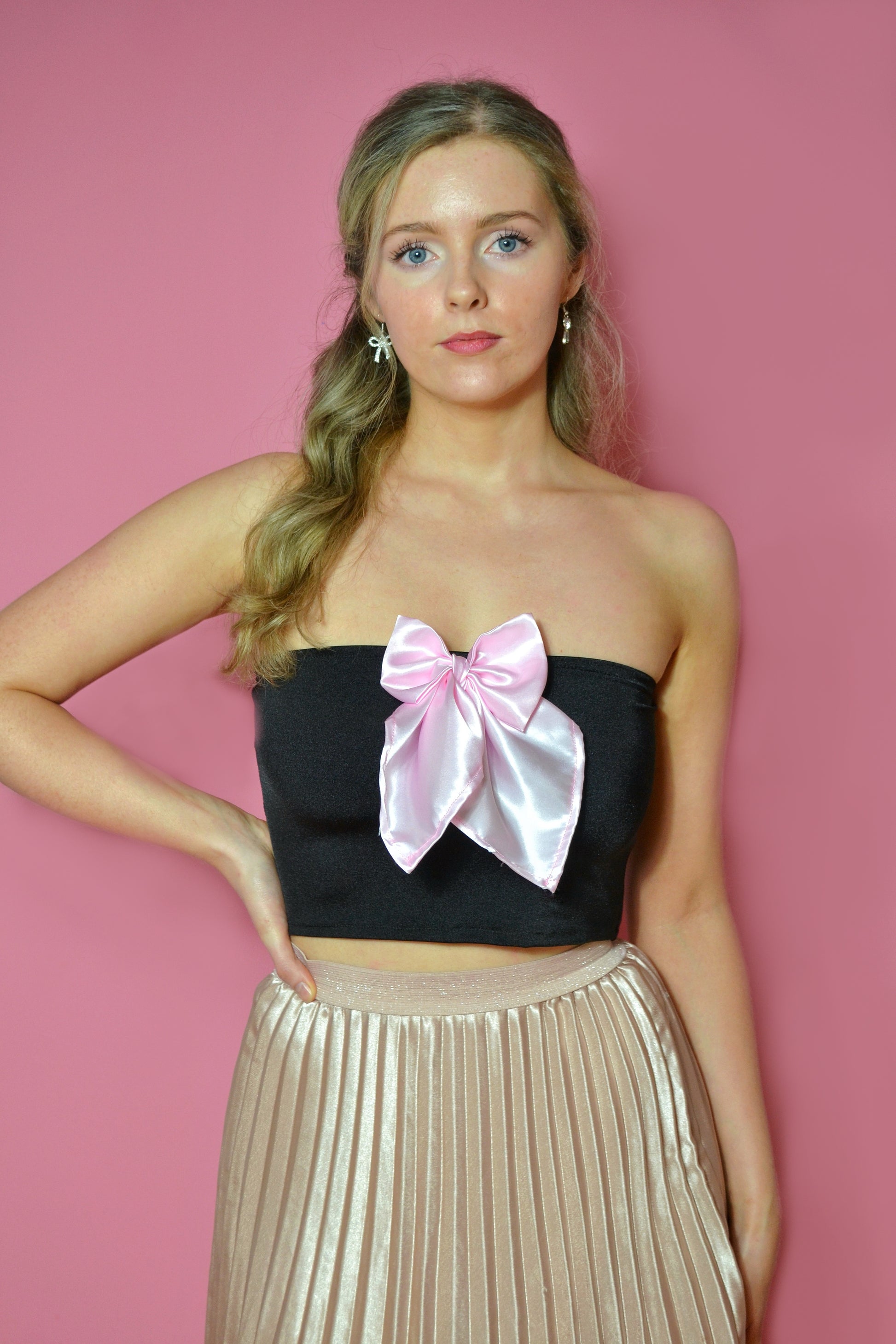 Woman wearing a black strapless top with a pink bow and a pale pink pleated skirt against a pink background