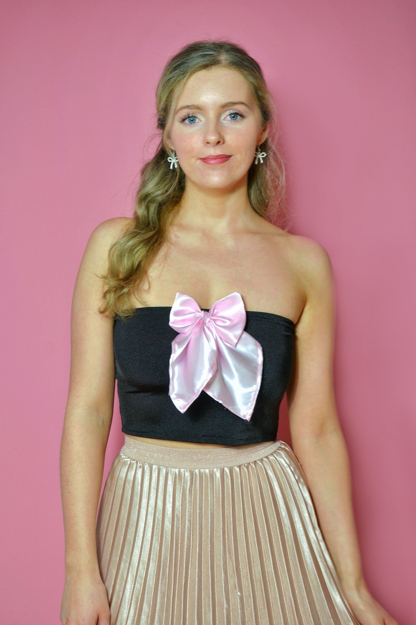 Woman wearing a black strapless top with a pink bow and a pale pink pleated skirt against a pink background