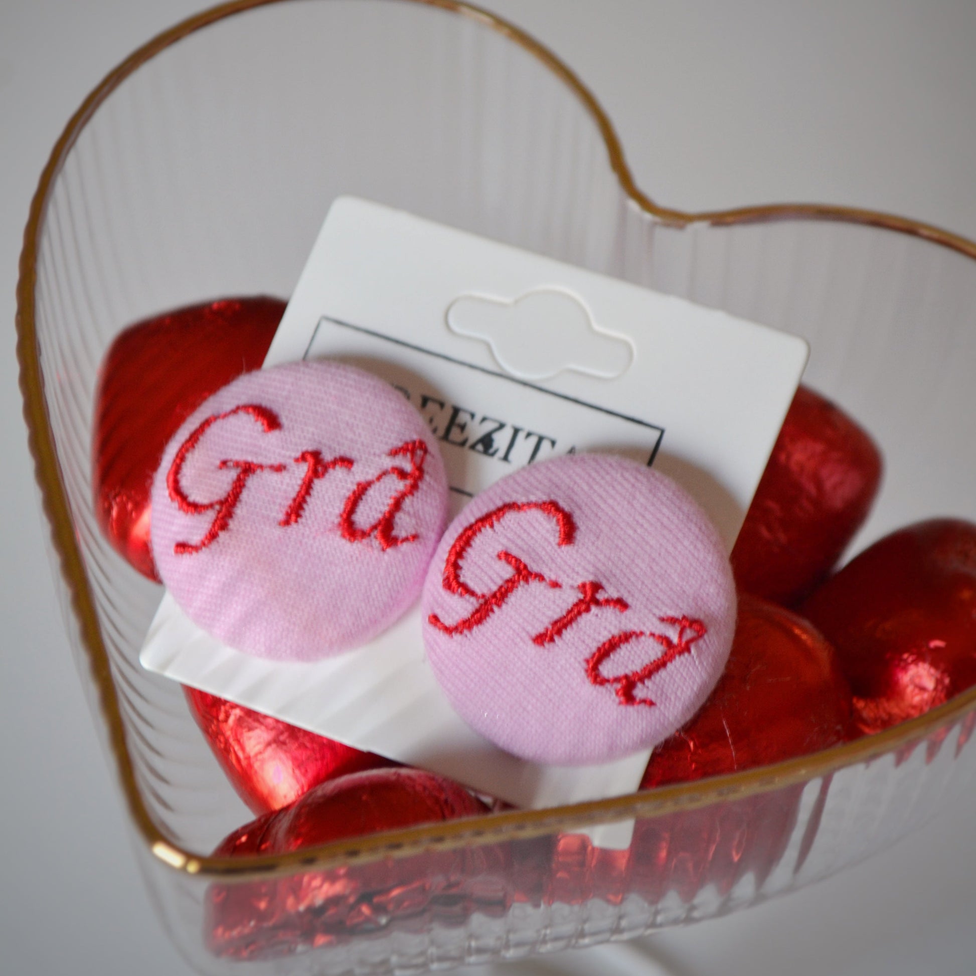 Pink earrings with 'Grá' text in a heart-shaped container with red candies.
