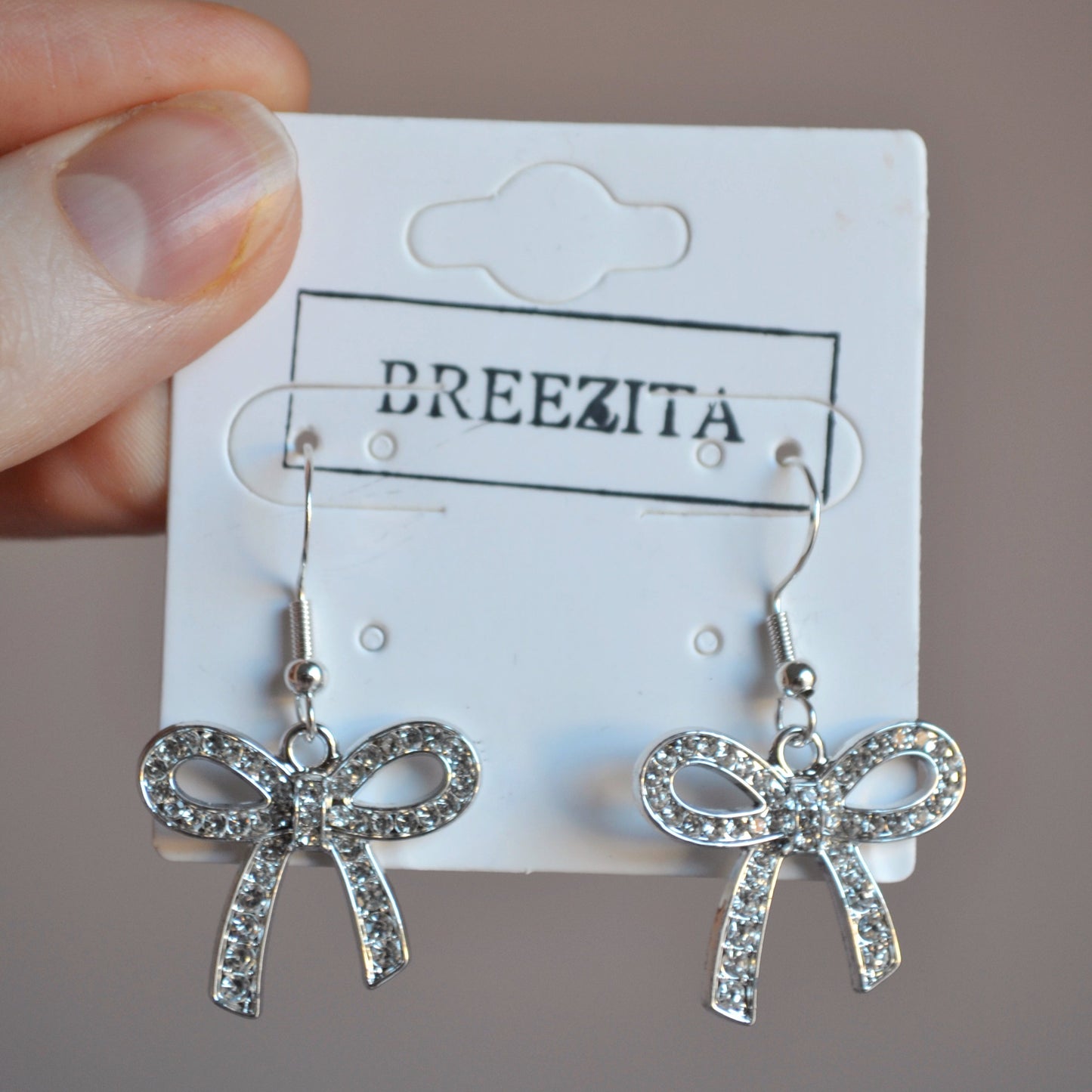 Silver bow-shaped earrings on a Breezita branded card held by a hand.