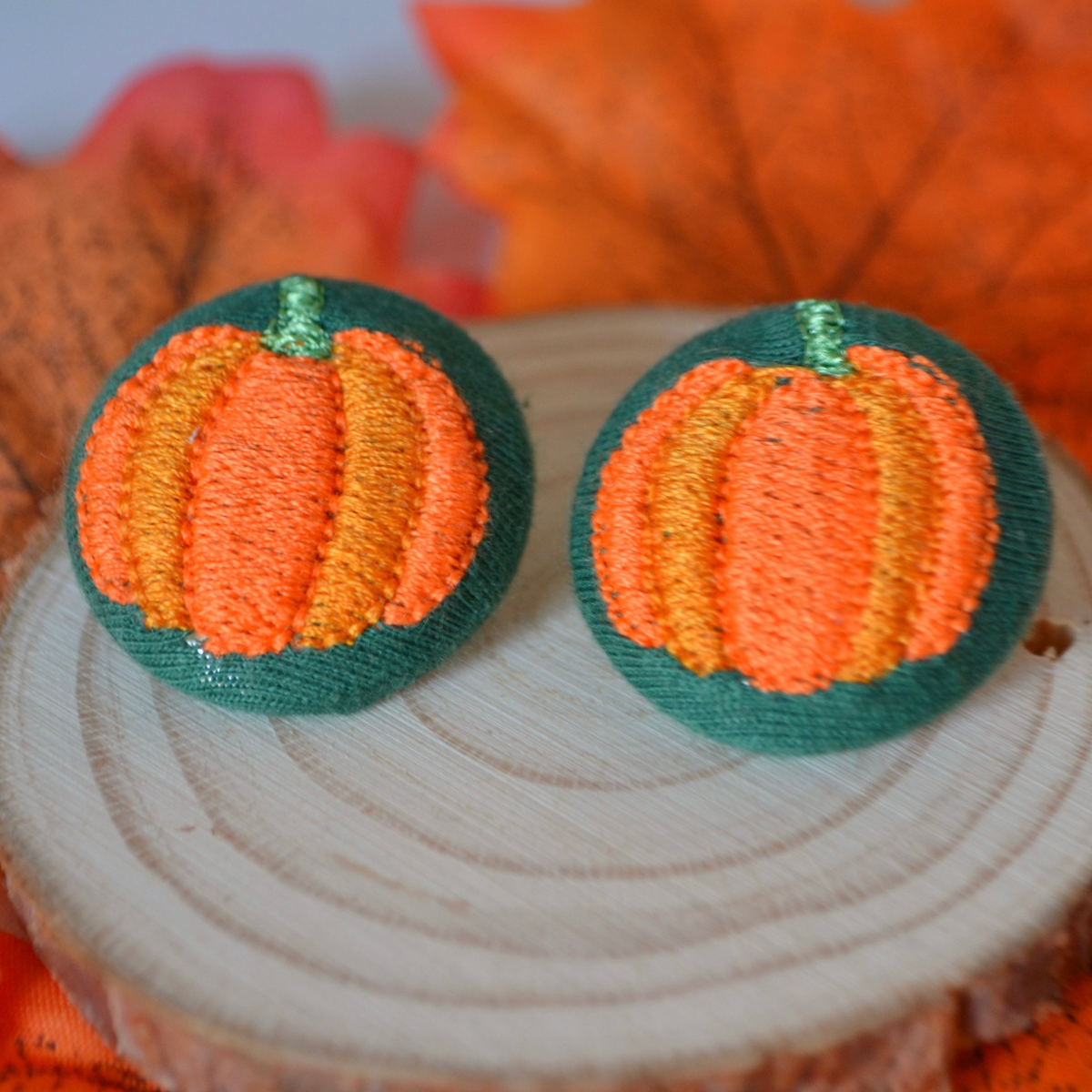Bottle-green fabric button earrings with orange pumpkin embroidery on a wooden display.