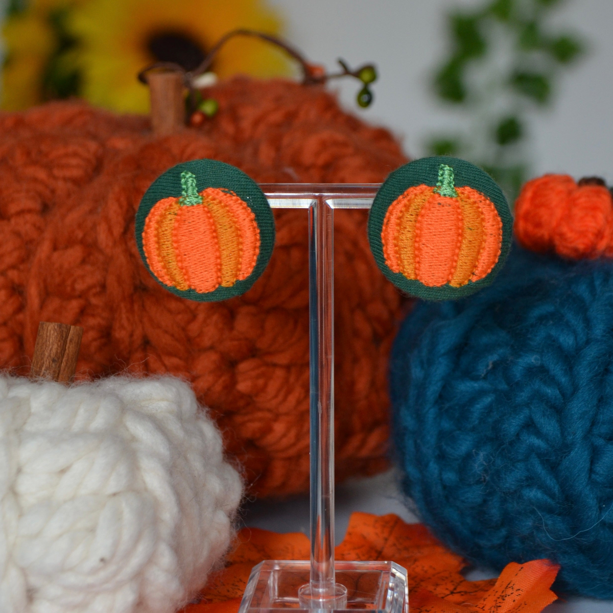 Orange embroidered pumpkin earrings on bottle-green fabric, displayed on an earring stand.