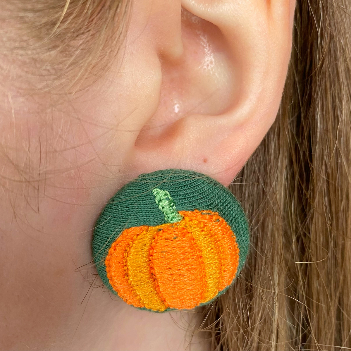 Bottle-green embroidered pumpkin button earring shown worn on an ear.