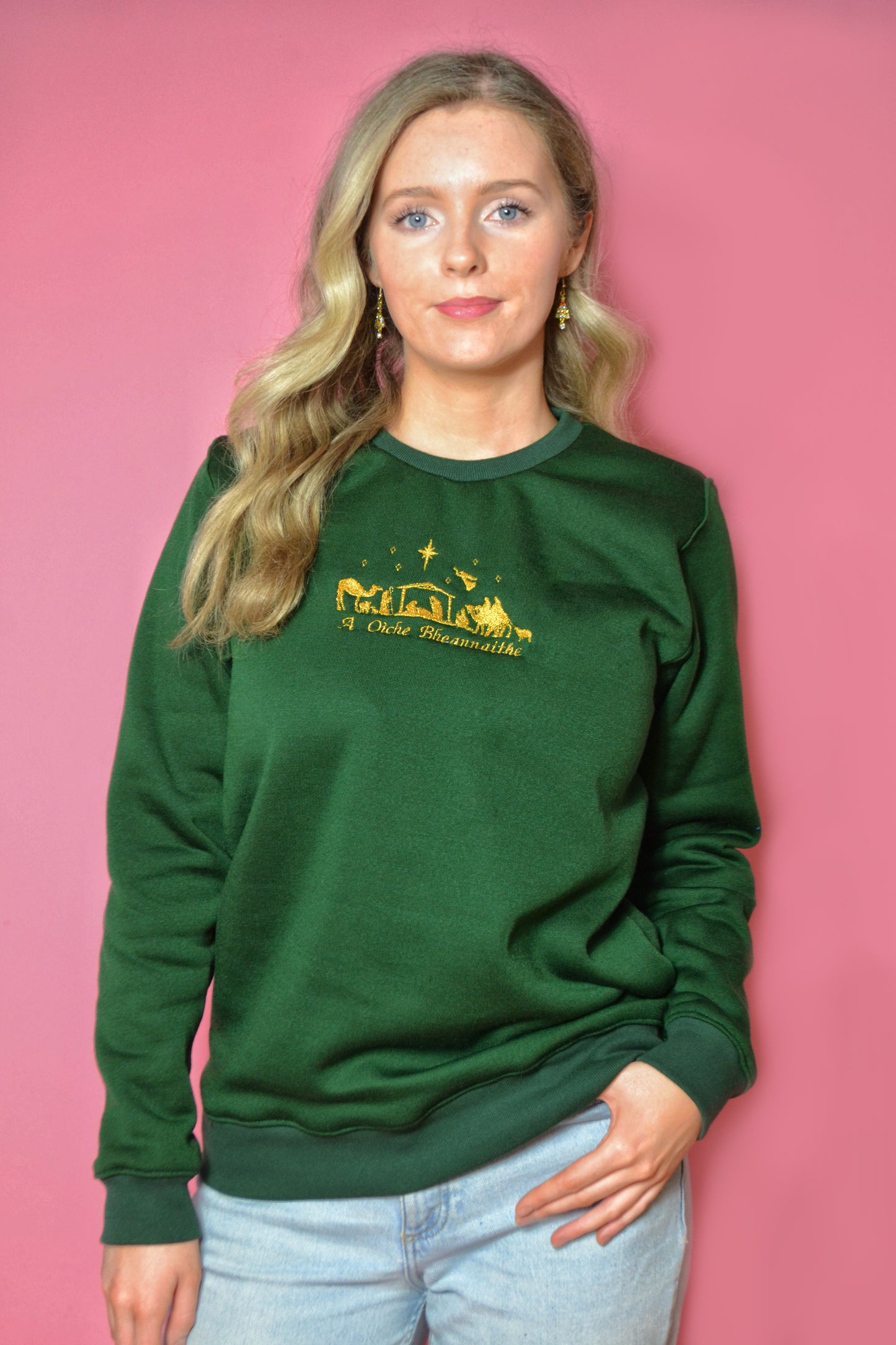 Woman wearing a green sweatshirt with Irish and the nativity embroidered onto it against a pink background