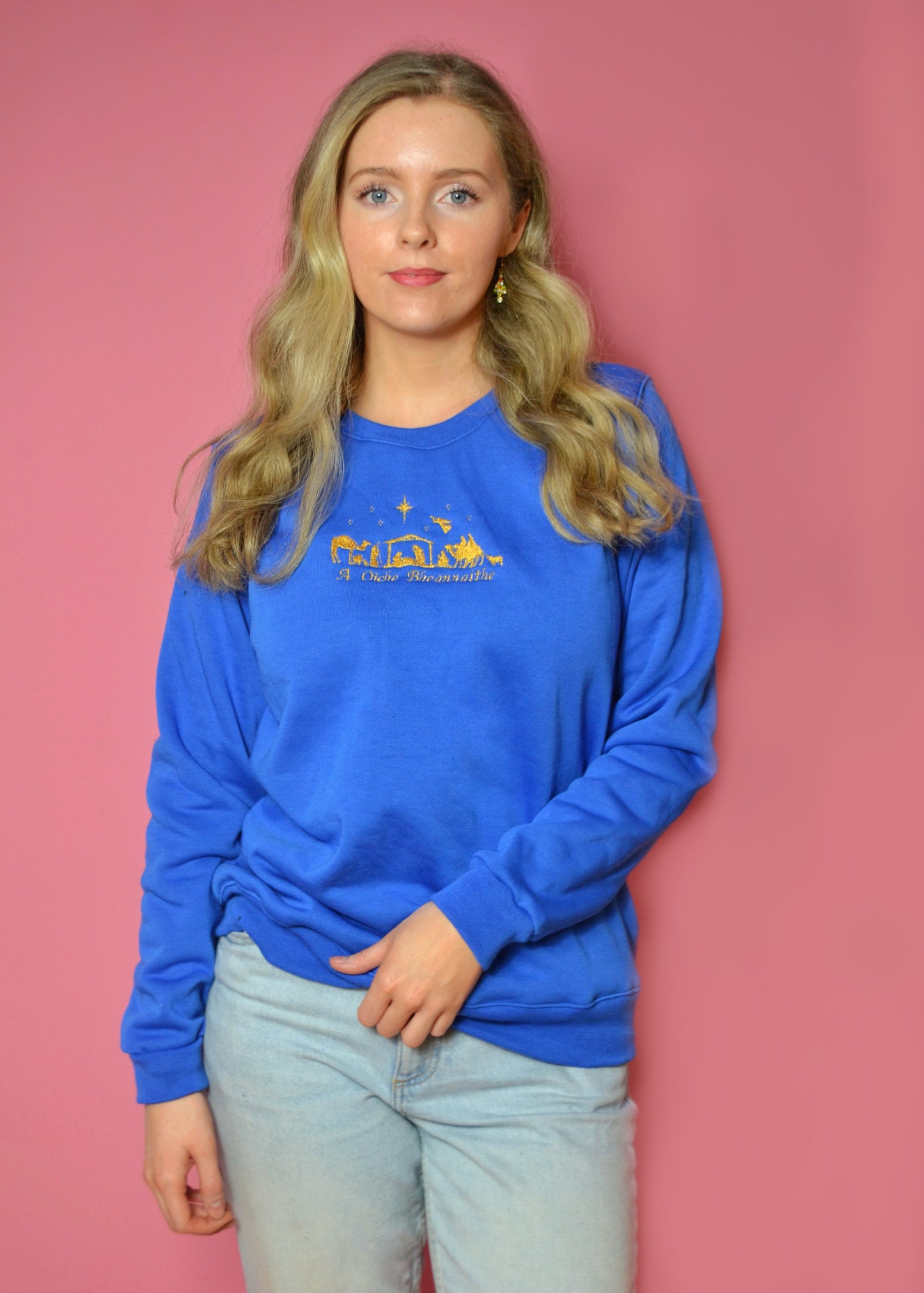 Woman wearing a blue sweatshirt with Irish and the nativity embroidered onto it against a pink background