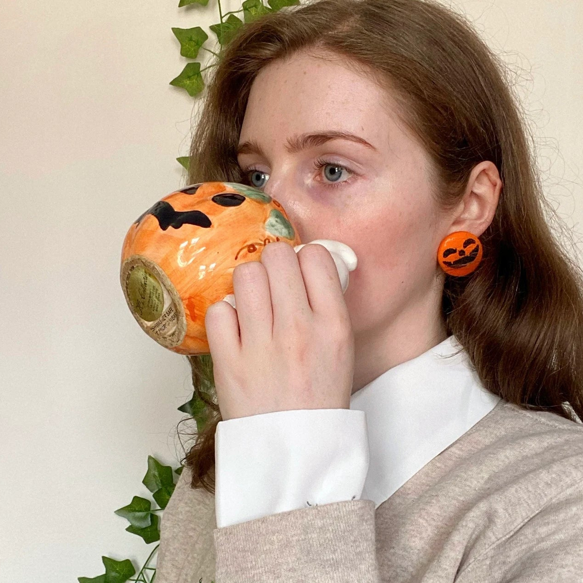 Woman wearing orange pumpkin face earrings while drinking from Halloween-themed mug, autumn cozy vibe.