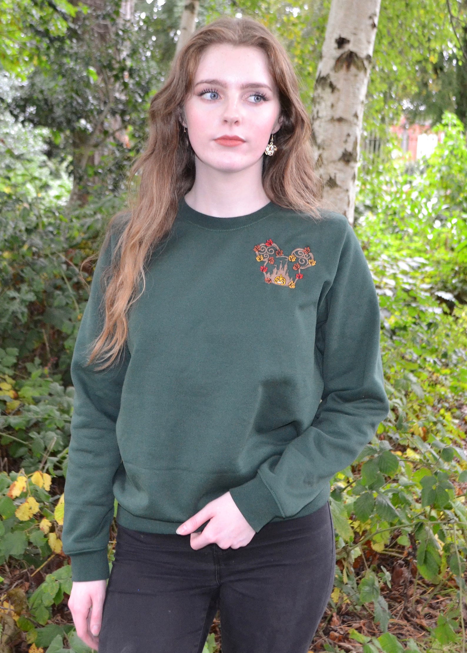 Female model styling bottle-green sweatshirt and black jeans; woodland pumpkin-leaf chest motif, cosy autumn streetwear